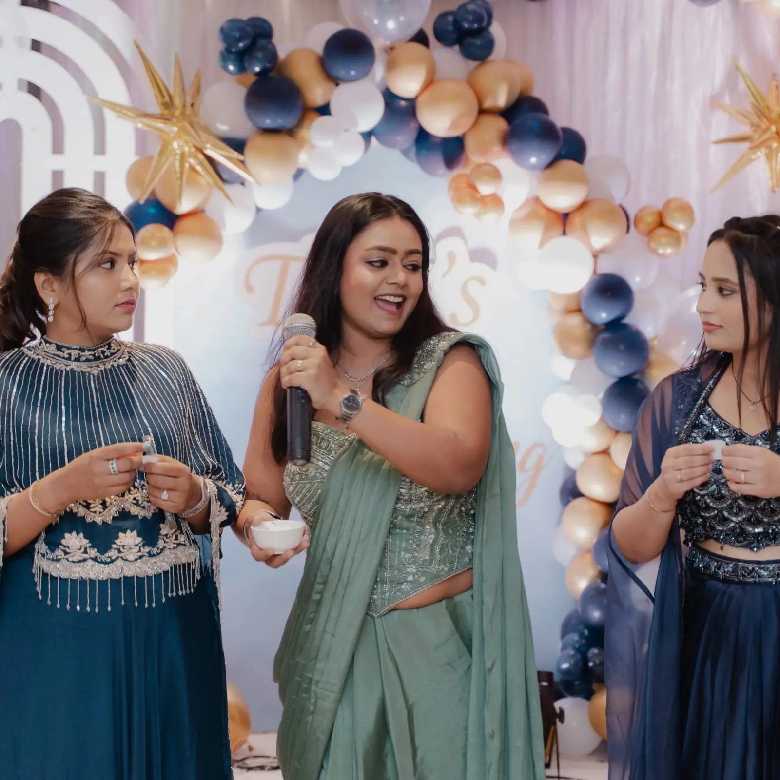 Palak Shah with a microphone between two guests in navy outfits at a celebration under a navy, gold and white balloon arch