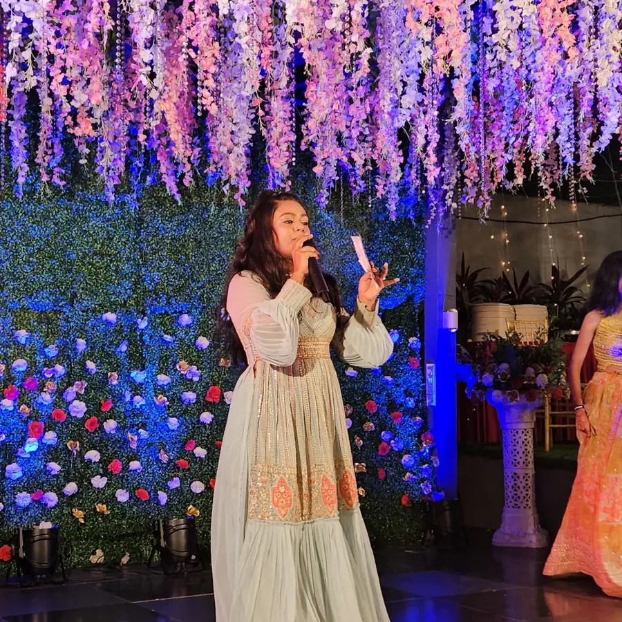 Palak Shah in a pale mint embroidered anarkali, holding a microphone and a cue card on an evening stage beneath a canopy of hanging wisteria flowers and a blue-lit flower wall