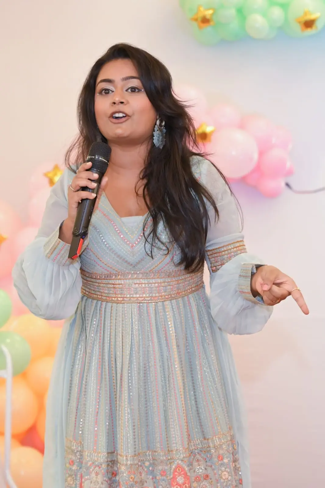 Palak Shah in a powder-blue embroidered anarkali with a microphone, pointing towards the guests in front of pastel pink, green and orange balloons