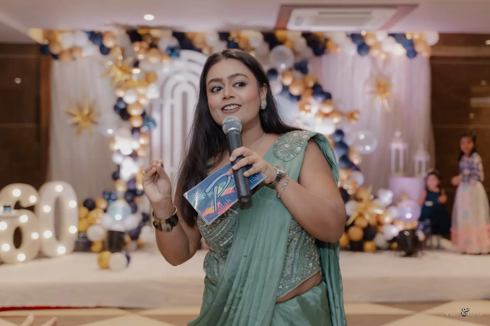 Palak Shah smiling with a microphone and cue card, hosting a birthday celebration beside balloon numbers spelling 60