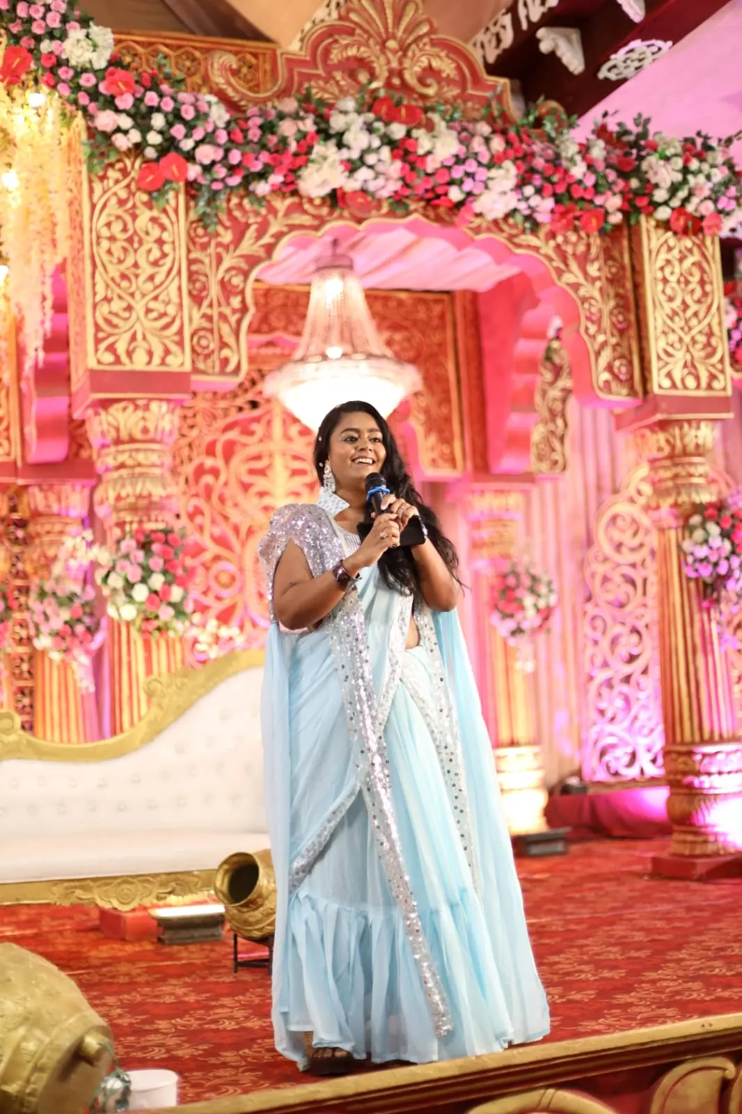 Palak Shah in a pale blue saree with a microphone, anchoring from an ornate pink and gold wedding stage decorated with flowers