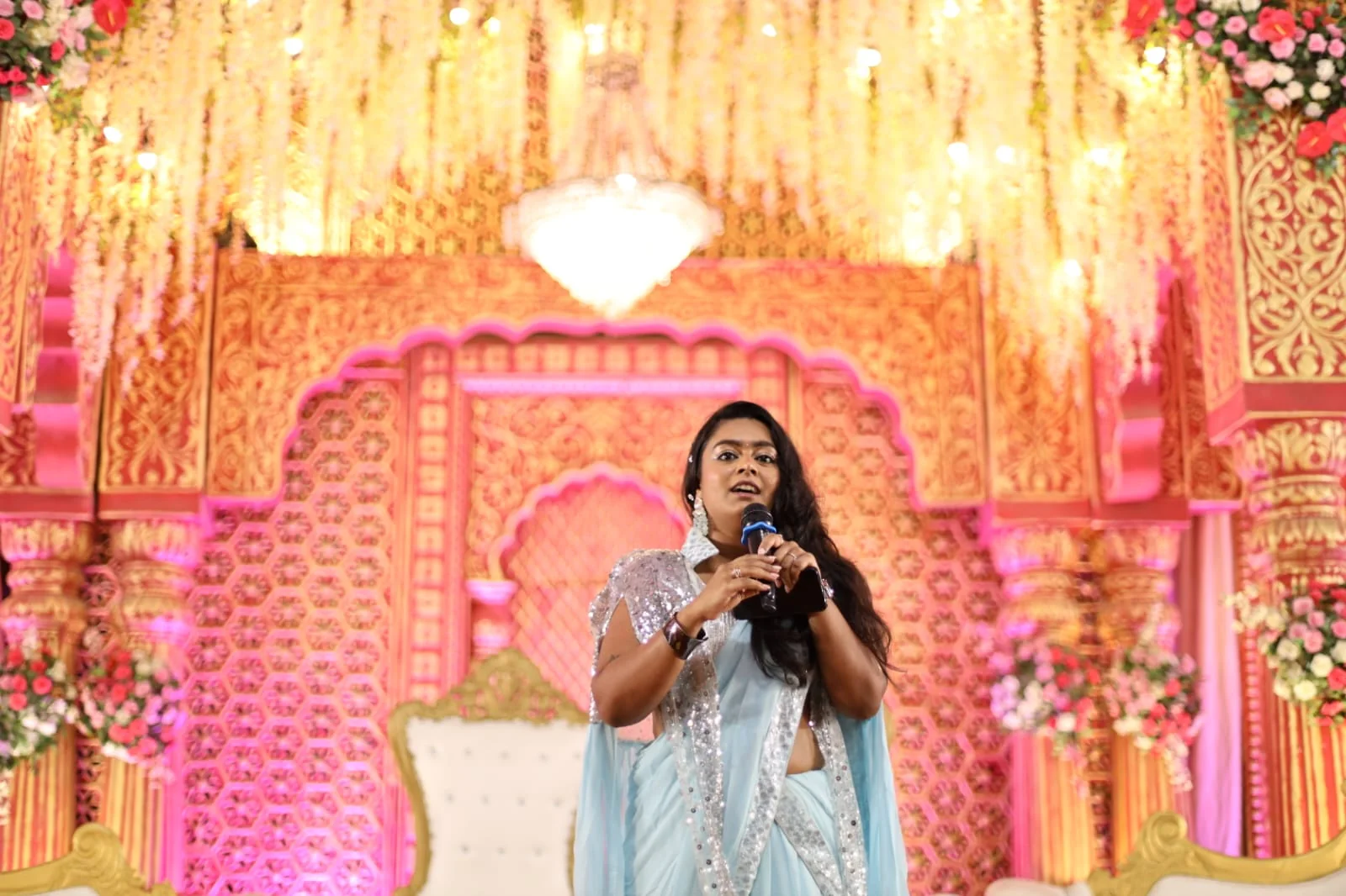 Palak Shah speaking into a microphone in front of a carved pink and gold wedding stage with a crystal chandelier and floral curtains