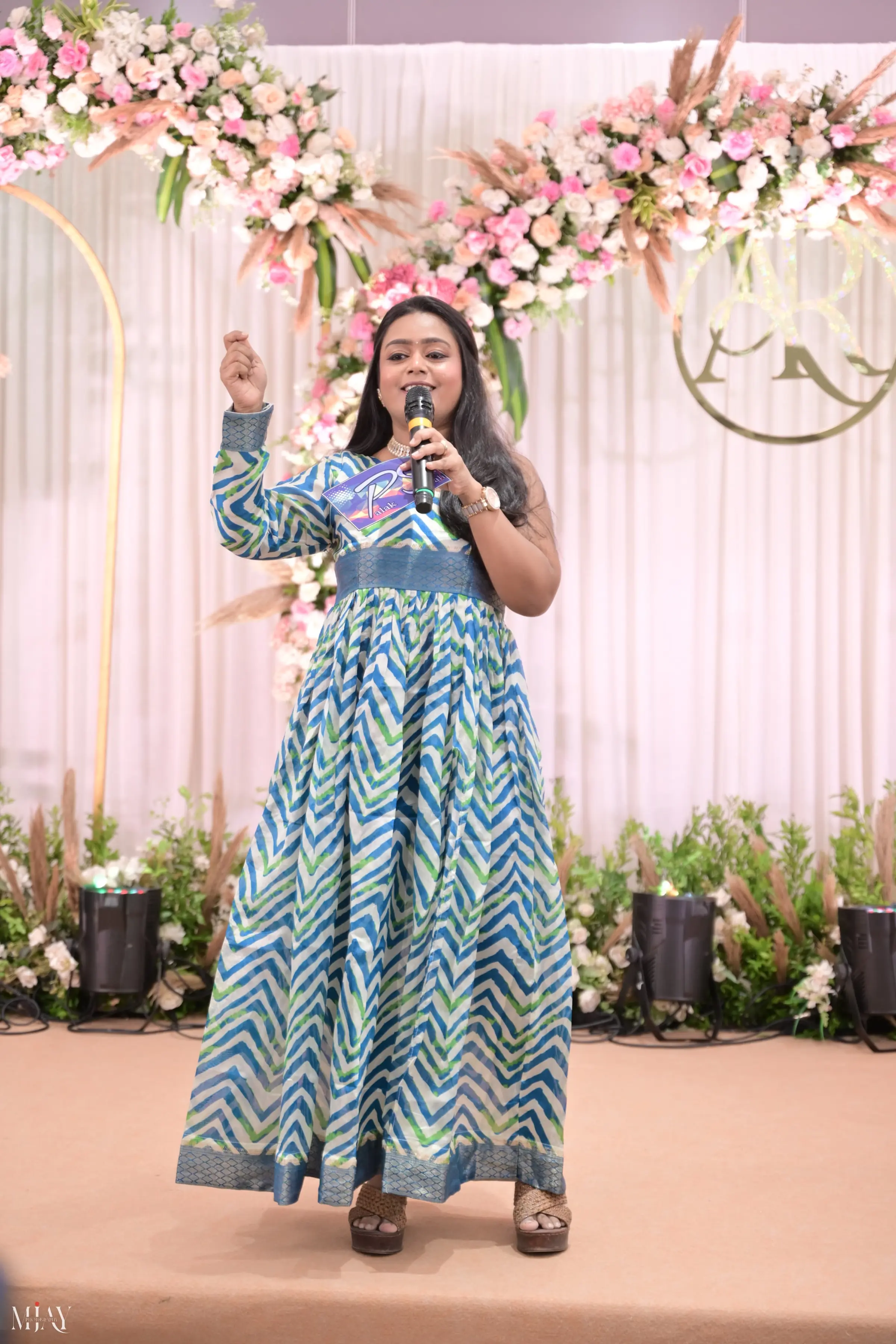 Palak Shah on stage with a microphone, one hand raised, in a blue and green chevron-print gown before a pink floral backdrop