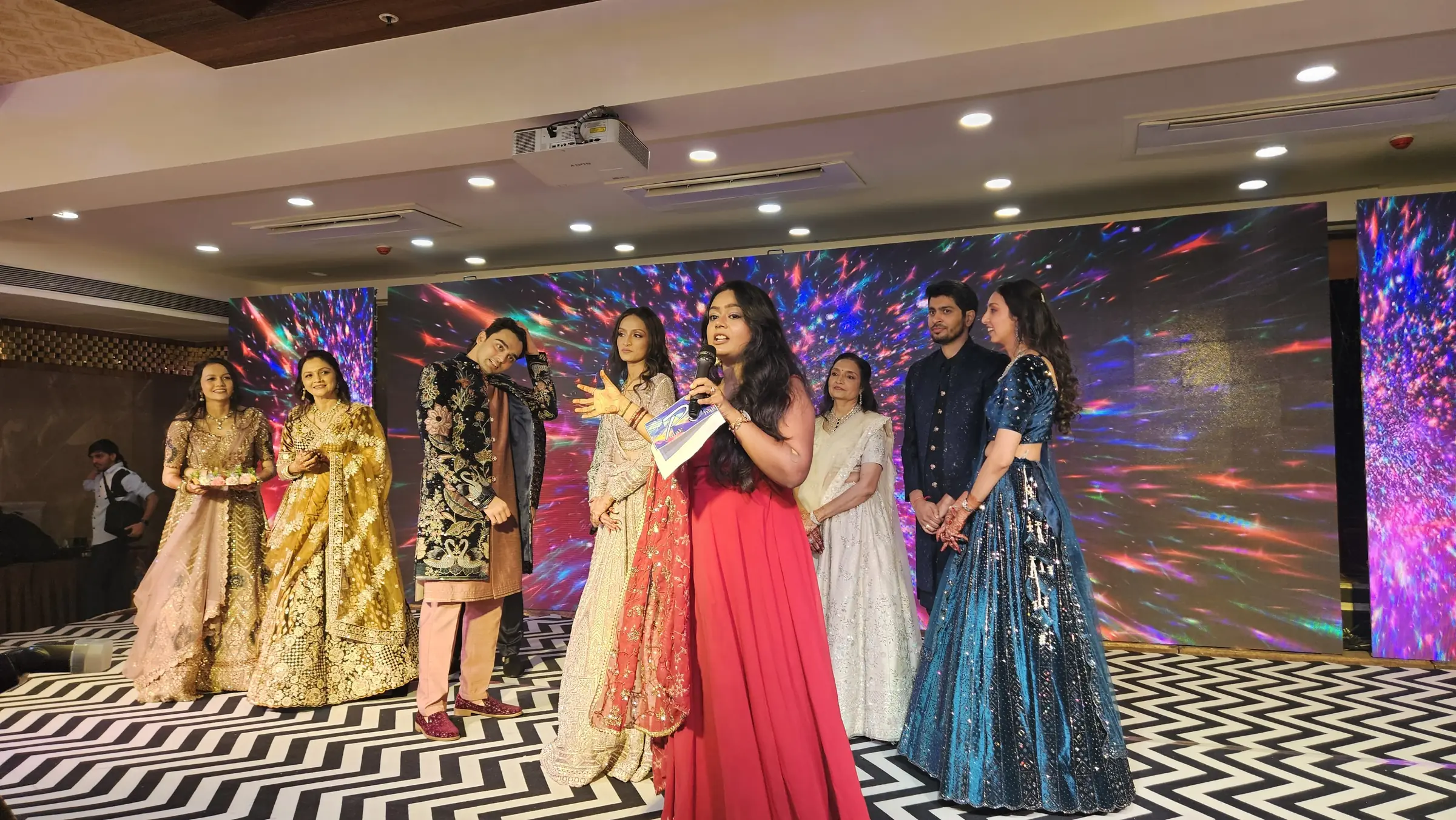 Palak Shah in a red gown, microphone in hand, hosting on stage beside a family in festive outfits, with a colourful light backdrop