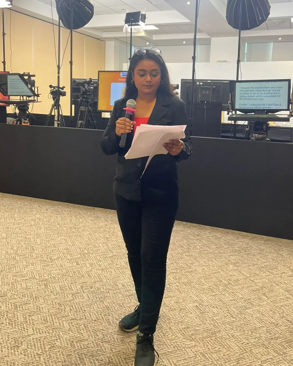 Palak Shah in a black blazer reading from her script with a microphone at a corporate AGM, with studio lights, cameras and teleprompters behind her