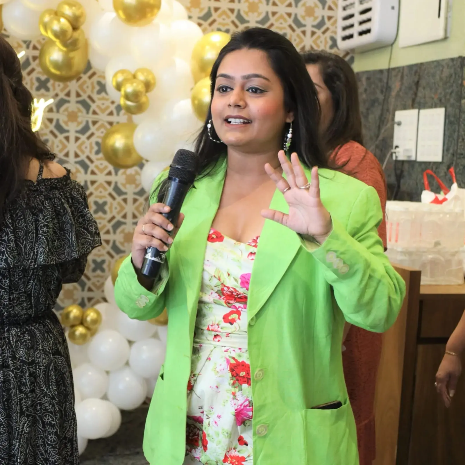 Palak Shah in a lime-green blazer over a floral dress, microphone in hand and one hand raised, hosting a celebration beside white and gold balloons